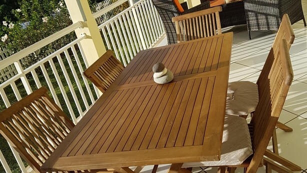 Uma mesa e cadeiras de madeira numa varanda com guarda-corpo branco, um sofá de vime e vista para o verde e edifícios.