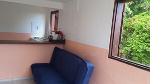 Ein Zimmer mit blauem Sofa, einer Küchenzeile mit Spüle und Kochutensilien sowie einem Fenster mit Blick auf Grünfläche.