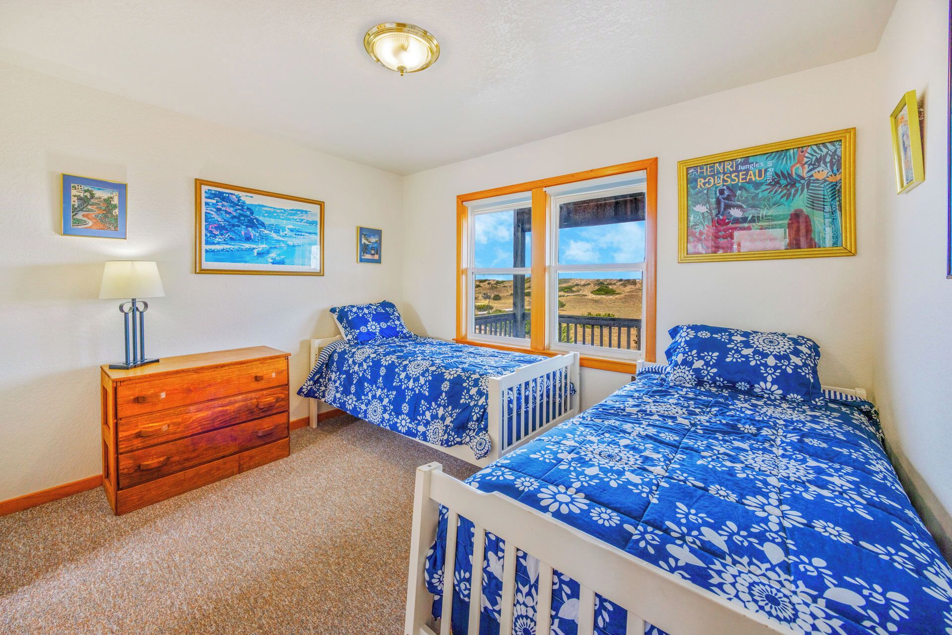 A bedroom with two beds, a wooden dresser, a lamp, and a window with a view.