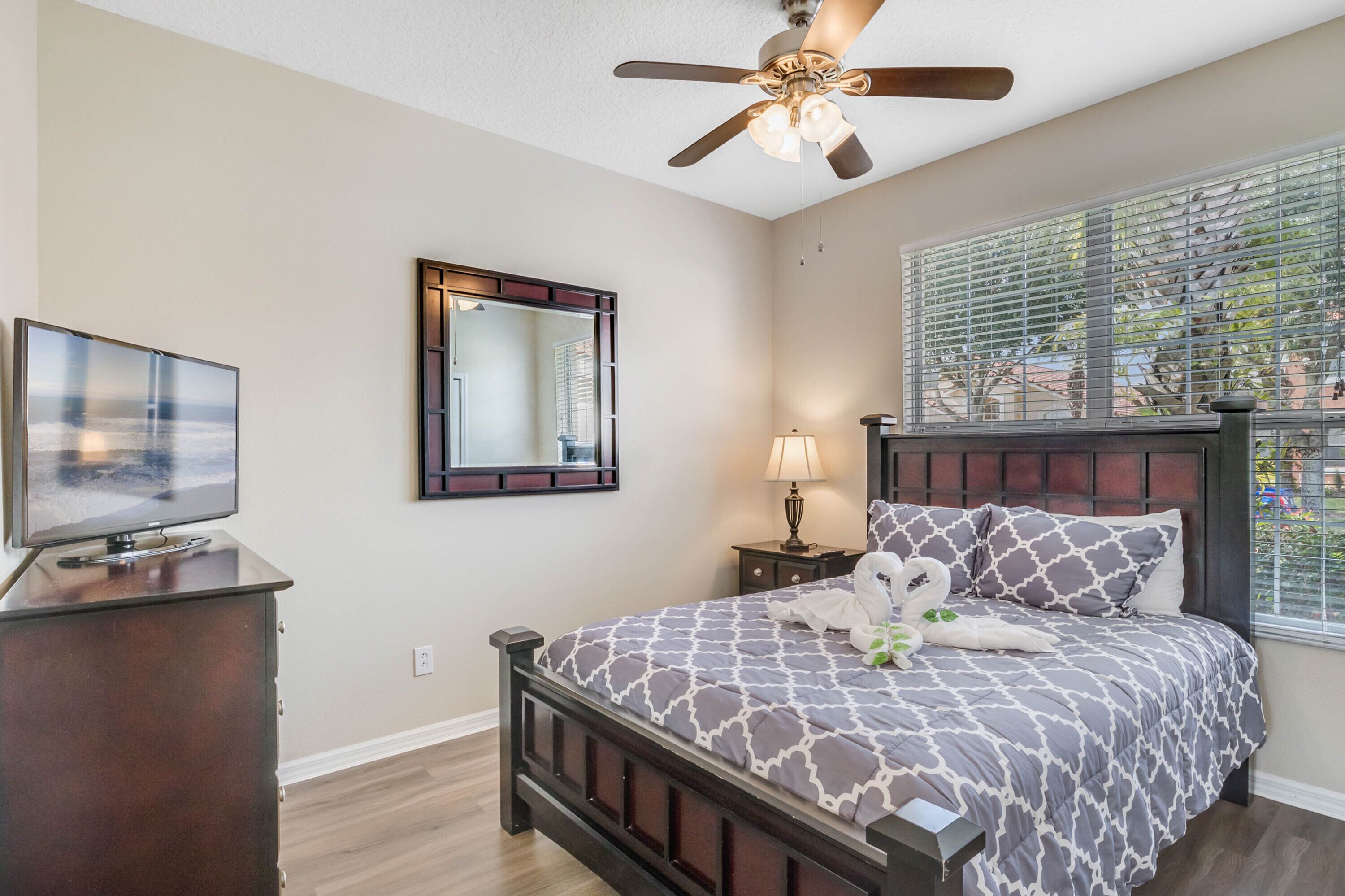 A bedroom with a bed, a television, a ceiling fan, a window with blinds, and a nightstand with a lamp.