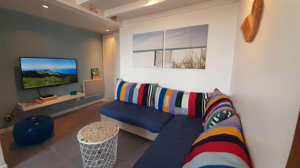 Ein modernes Wohnzimmer mit Flachbildfernseher, einer Couch mit bunten Kissen, einem kleinen runden Couchtisch und einer Wandregal.