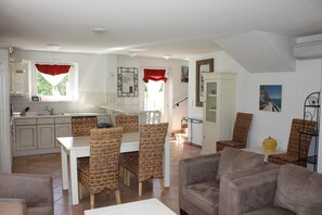 Un vaste espace de vie comprenant une table à manger, des chaises en osier, un canapé et une cuisine avec des meubles blancs et une fenêtre ornée de rideaux rouges.