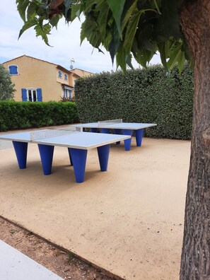 Des tables de ping-pong installées à l’extérieur, avec des pieds bleus, posées sur une surface sablonneuse, entourées de verdure et d’un bâtiment aux volets bleus.