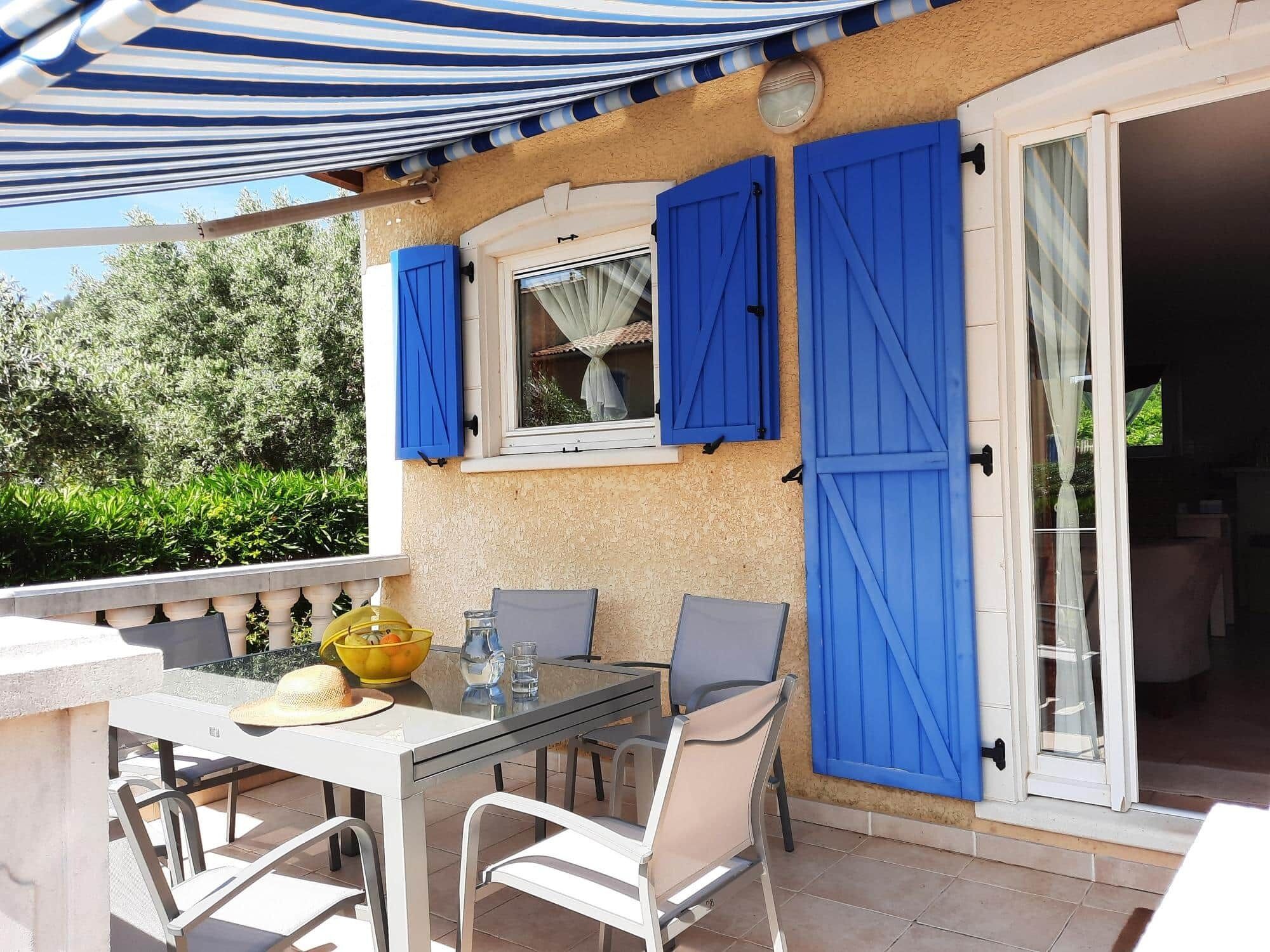 Une terrasse extérieure avec une table et des chaises, une fenêtre avec des volets bleus et une porte donnant sur un espace intérieur.