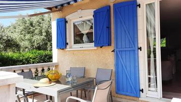 Une terrasse extérieure avec une table et des chaises, une fenêtre avec des volets bleus et une porte donnant sur un espace intérieur.