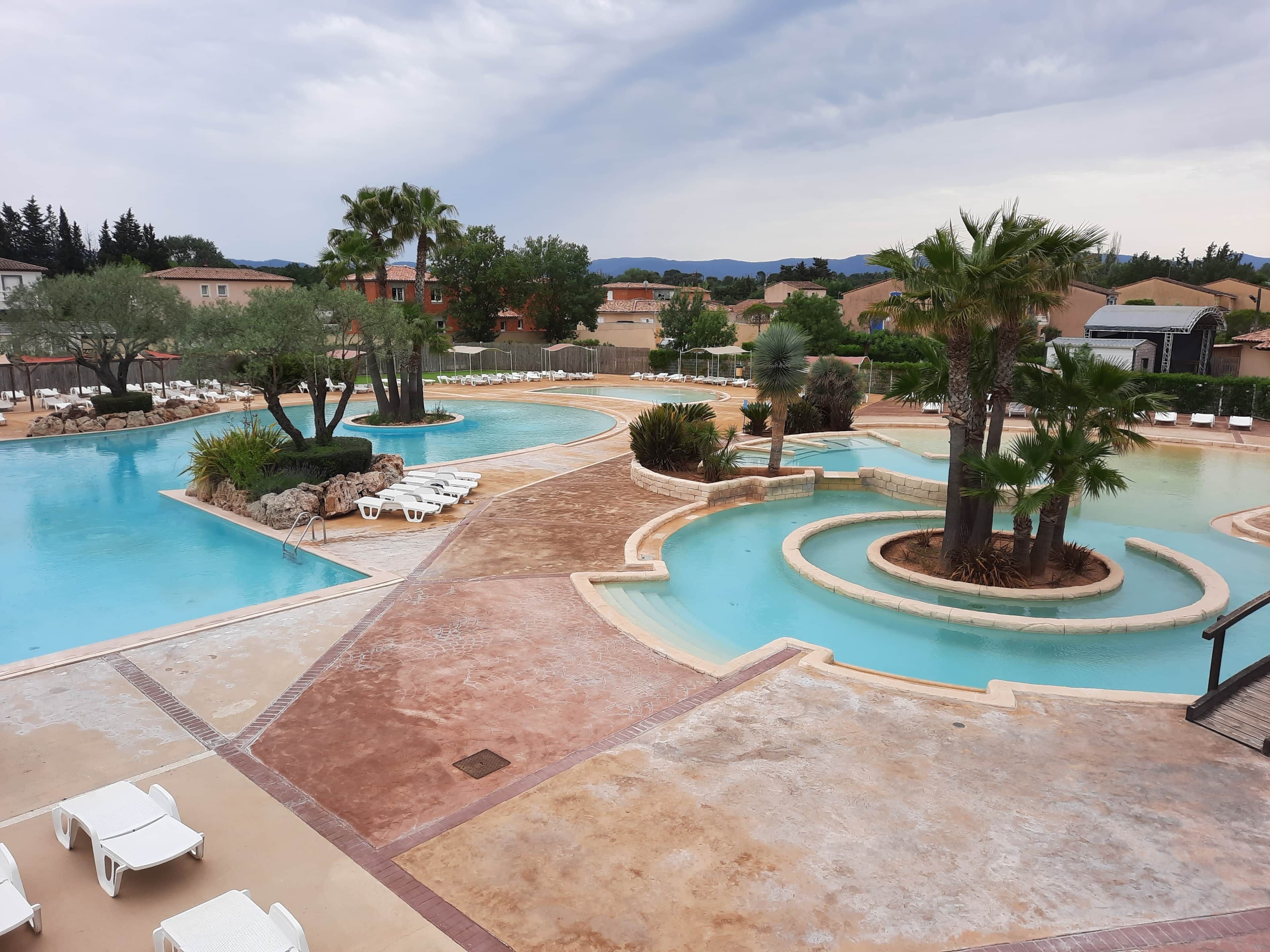 Un espace piscine dans un complexe hôtelier, comprenant plusieurs piscines, des chaises longues et des palmiers.