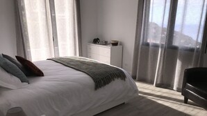 Een moderne slaapkamer met een groot bed, nachtkastje, dressoir en uitzicht op het landschap door dunne gordijnen.
