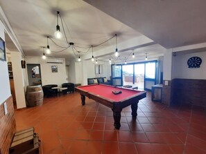 Une table de billard avec un éclairage suspendu, un jeu de fléchettes et une vue sur l’extérieur.