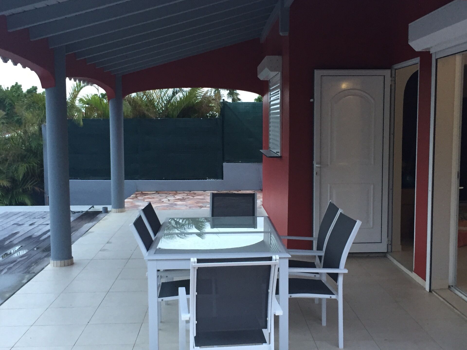 A covered patio area with a table and chairs, a door, and a view of a green fence and trees.