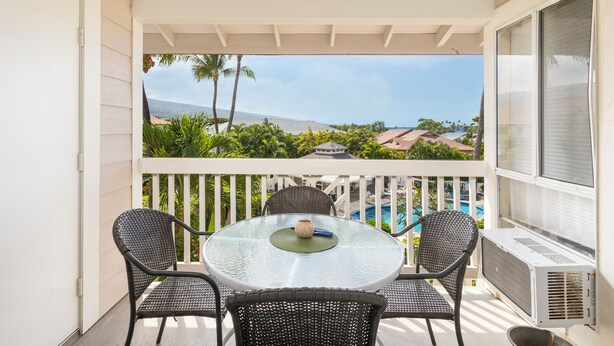 A balcony with a round glass table, two wicker chairs, and a view of a swimming pool and palm trees. - 🌴Lovely quiet location partial ocean views.This complex requires a 30 day minimum stay.🌴 (Kailua Kona)