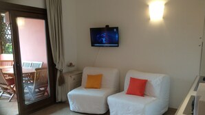 Ein Zimmer mit zwei weißen Sofas, eines mit einem orangefarbenen Kissen und das andere mit einem roten Kissen, einem am Wand montierten Flachbildfernseher und einer Glastür, die zu einem Außenbereich mit Tisch und Stühlen führt.