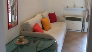 Ein Wohnzimmer mit einem Glastisch, einem beige farbenen Sofa mit bunten Kissen, einem weißen Heizkörper und zwei an der Wand hängenden Bildern.