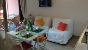 Ein Essbereich mit Tisch für zwei Personen, eine weiße Couch mit orangenen Kissen, ein Flachbildfernseher an der Wand und ein Fenster mit Vorhängen.