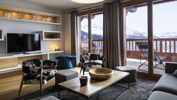 A modern living room with a large flat-screen TV, a wooden coffee table, and a sofa. There is a balcony with a view of snow-capped mountains. - Appart 130m², full services 4 *, 4 bedrooms, 4 bathrooms / d, south balcony and 2 parking spaces (Montvalezan)