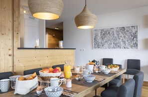 A breakfast table set with croissants, fruits, and a glass of orange juice in a modern kitchen with wooden accents and hanging pendant lights.