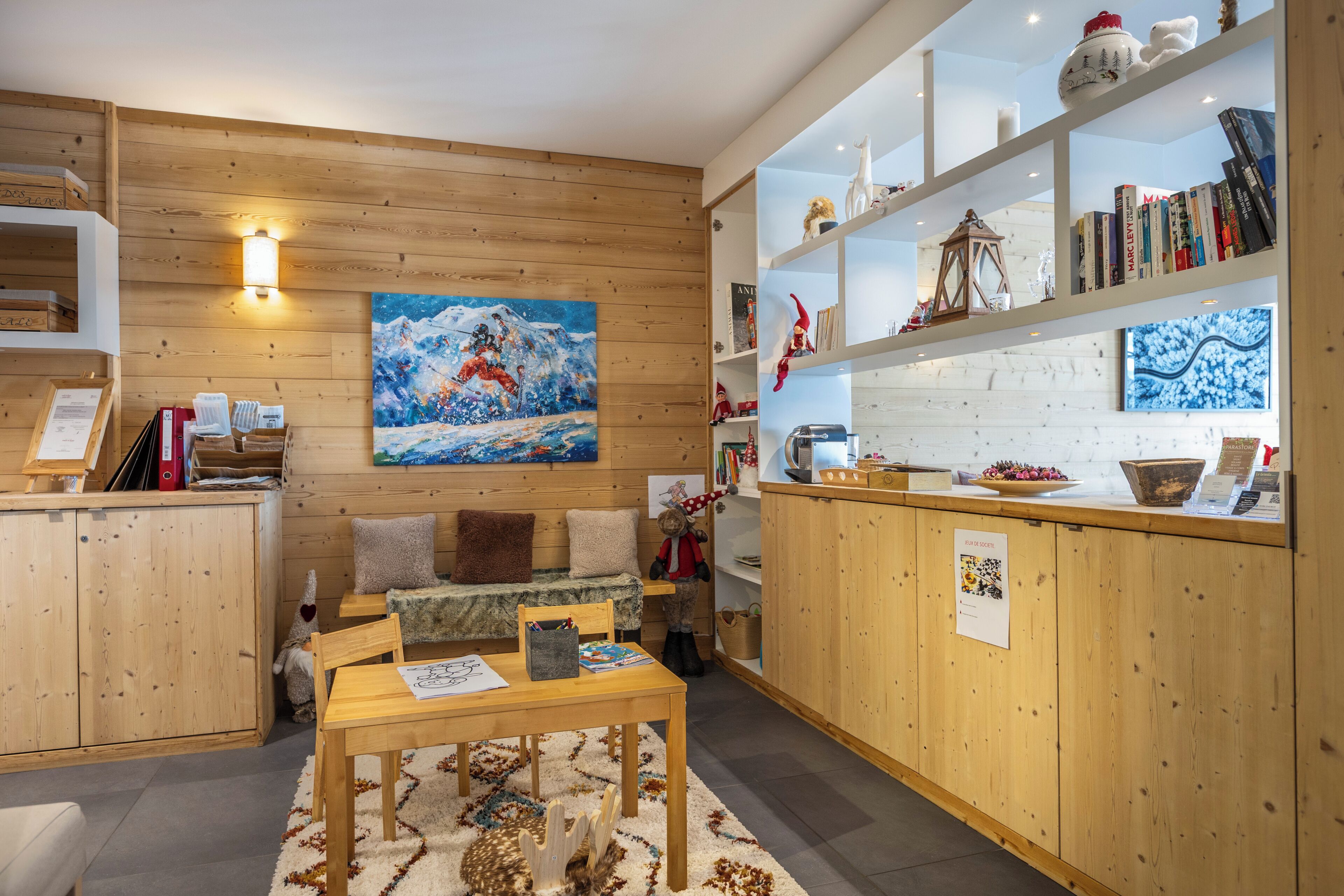 Een knus vertrek met houten wanden, een schilderij van een besneeuwde berg, een houten tafel en een boekenkast met boeken en decoratieve objecten.