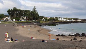 Ein Strand mit badenden Personen, einer felsigen Küstenlinie und Häusern im Hintergrund.