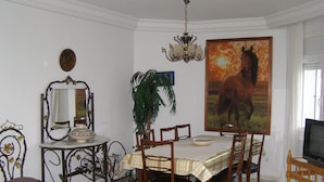 Un comedor con una mesa dispuesta para cuatro personas, rodeada de sillas de madera. Un cuadro grande de un caballo adorna la pared, y un candelabro cuelga del techo.