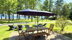 Een terras buiten met een houten tafel en stoelen, een grote parasol en uitzicht op het meer.