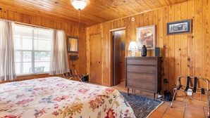 A room with wooden walls, a bed with a floral bedspread, a dresser, a lamp, and a framed picture on the wall. - River Ridge Cabin | Bandera Stay with River Access (Bandera)