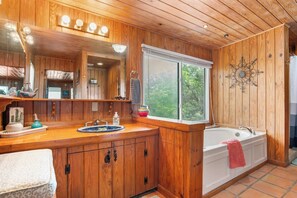 Un bany de fusta amb un mirall gran, un lavabo, una banyera i una finestra amb vista a vegetació.