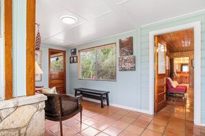 A hallway with a stone wall, wooden bench, and a wicker chair. There are framed pictures on the wall and a window with a view of trees. - River Ridge Cabin | Bandera Stay with River Access (Bandera)