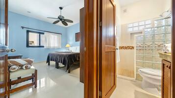 A hotel room with a bed, a desk, a chair, a ceiling fan, and a bathroom with a toilet and shower.