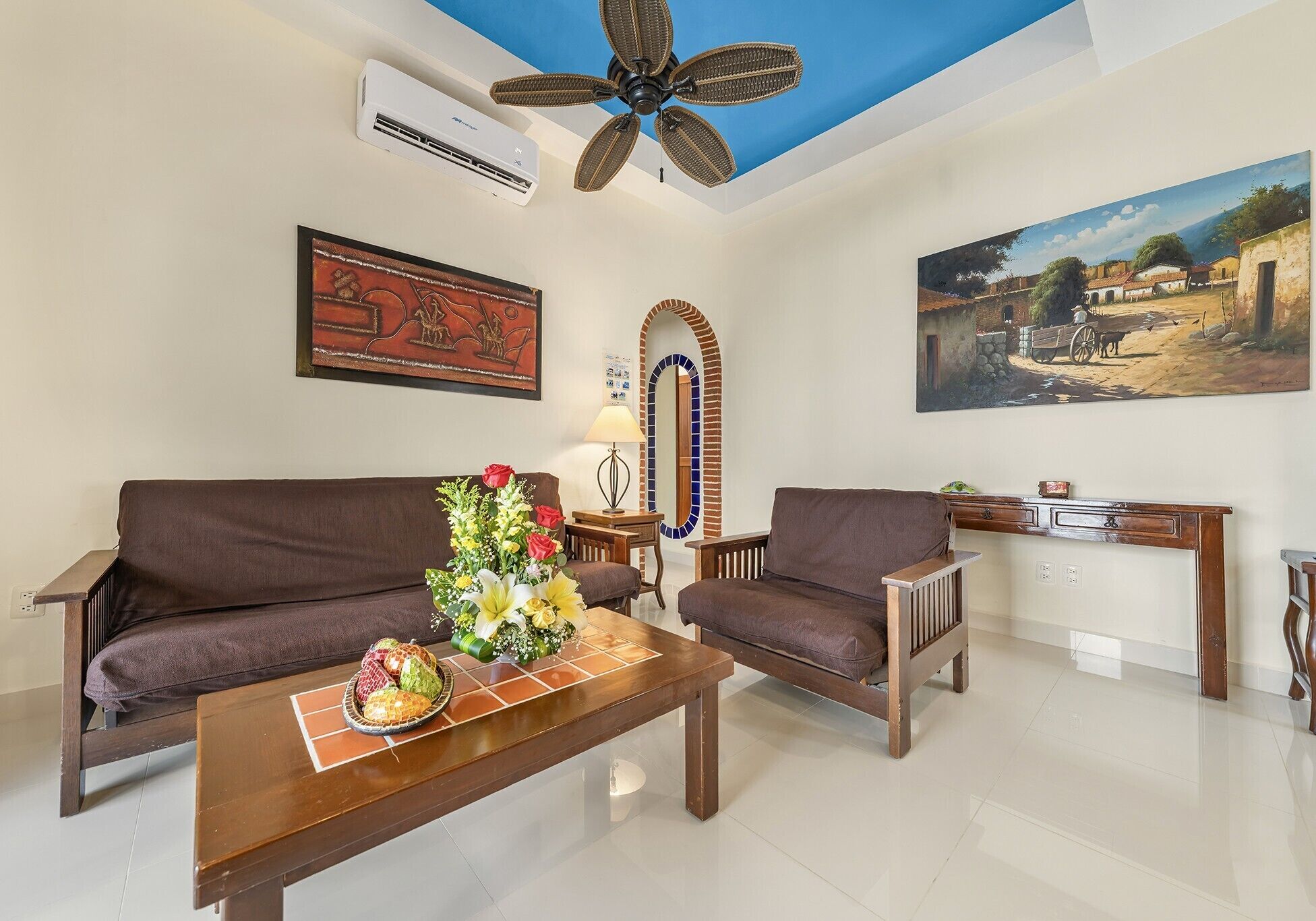 A living room with a brown sofa, a wooden coffee table with a floral centerpiece, a ceiling fan, an air conditioning unit, and a painting of a rural scene.