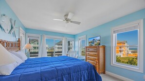 Ein Schlafzimmer mit Bett, Deckenventilator, Kommode und großem Fenster mit Blick auf Gebäude und Strand.