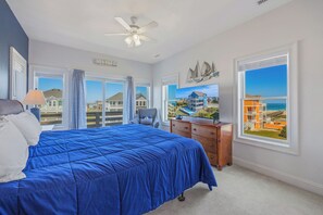 A bedroom with a bed, dresser, and large windows offering views of buildings and the beach. - Oceanview Dog Friendly Home w/ Private Pool, Hot Tub, Elevator, and Game Room (Rodanthe)