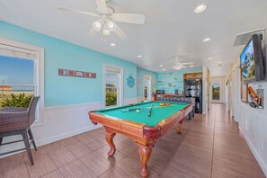 A pool table in a room with a ceiling fan, a view of the beach, and a kitchen area.