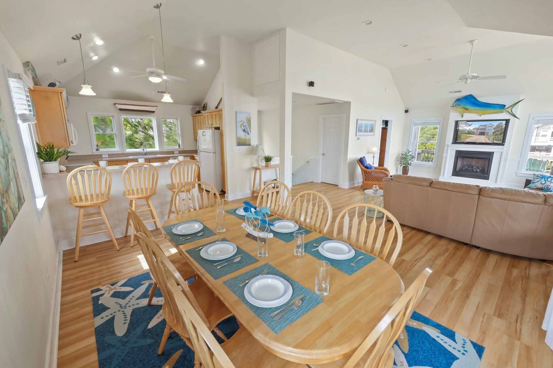 Une vaste salle à manger avec une longue table en bois, dressée pour six personnes, entourée de chaises en bois. On y trouve également un coin salon confortable avec un canapé et une cheminée.