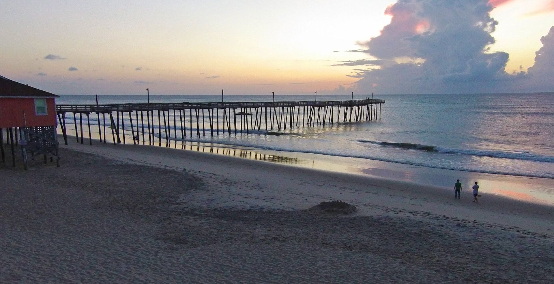 A beach with a wooden pier extending into the ocean, a small building on the left, and two people walking on the sand.