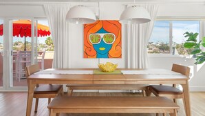 Un espace repas comprenant une table, des chaises, un tableau et une vue dégagée sur l’extérieur grâce à de grandes fenêtres.