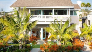 Une maison à deux étages avec un balcon, entourée de plantes tropicales et d’un petit jardin.