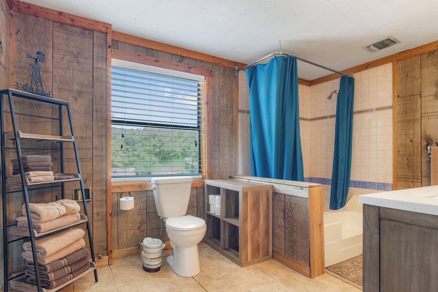 A bathroom with a toilet, a shelf with towels, a window with blinds, and a shower area.