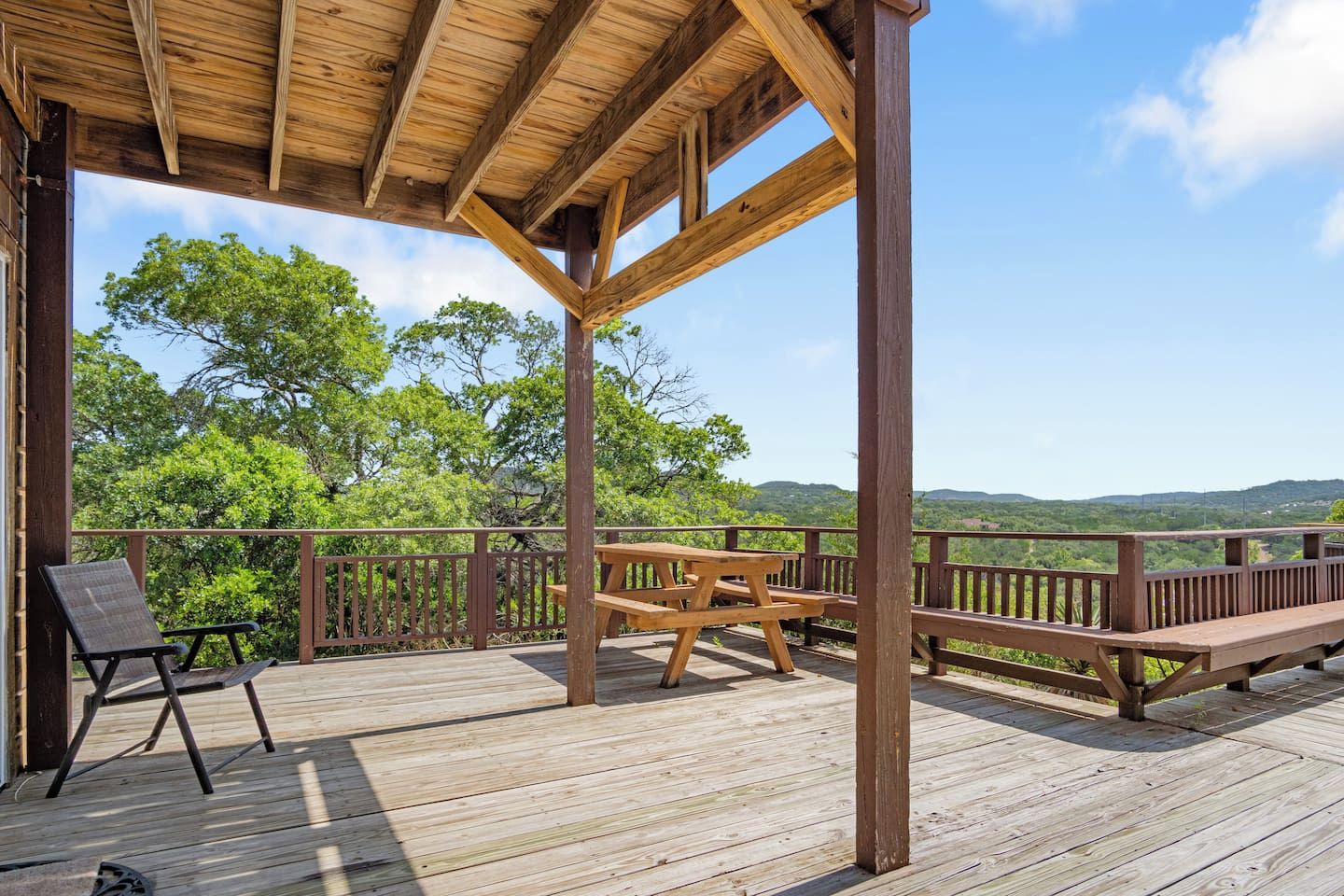 A wooden deck with a picnic table and chairs, surrounded by a railing with a view of green hills.