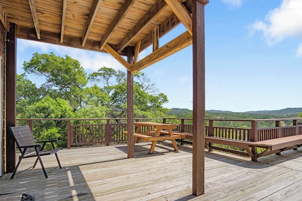 A wooden deck with a picnic table and chairs, surrounded by a railing with a view of green hills. - Rockin B Bluff | Hilltop 2BR Cabin with Hot Tub (Bandera)
