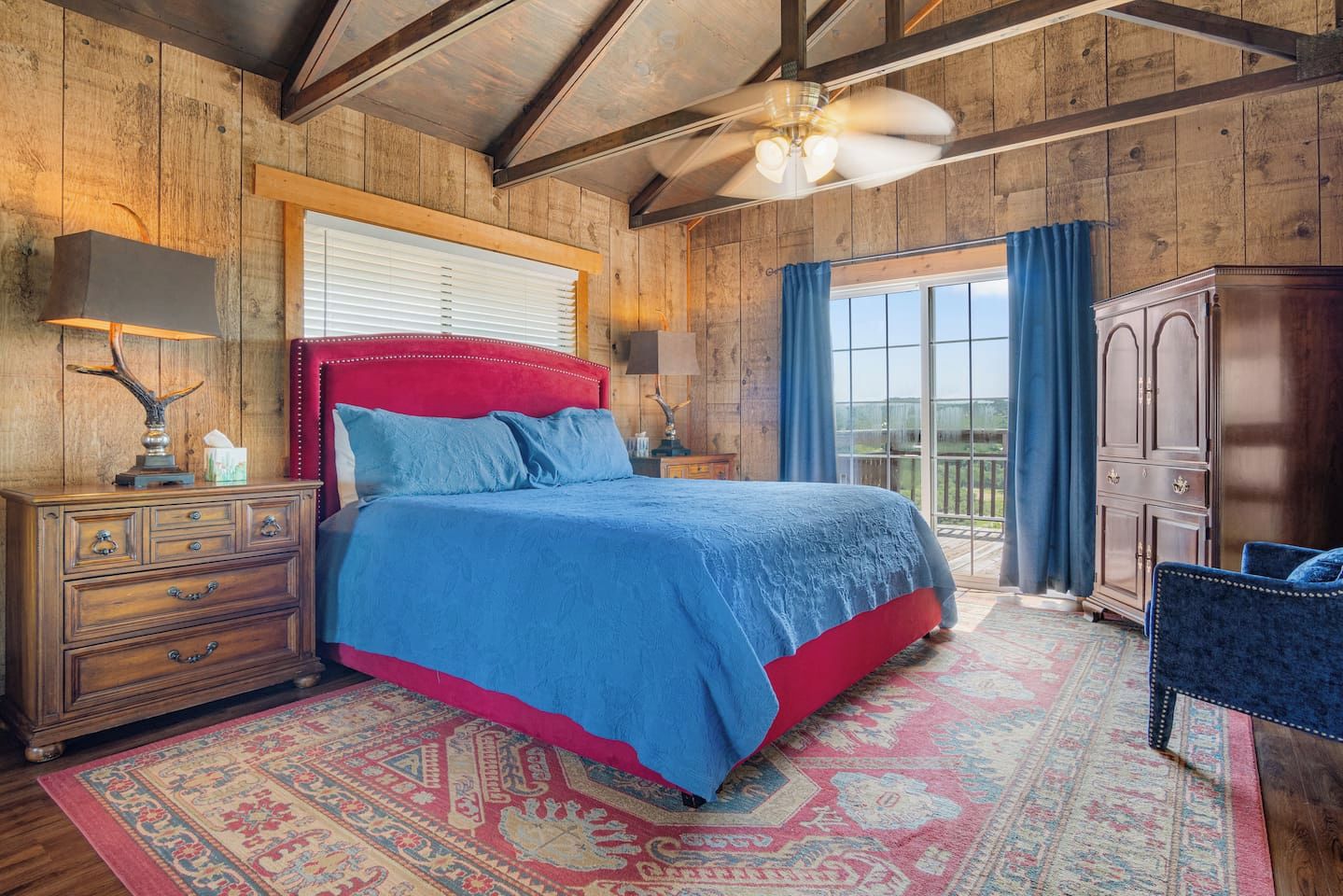 A bedroom with a large bed, wooden walls, a ceiling fan, a nightstand, a dresser, and a view through a window.