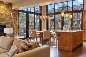 Ein geräumiges Wohnzimmer mit Steinwand, einem Esstisch mit Stühlen, einer Kücheninsel mit Spüle und großen Fenstern mit Blick auf eine Wohngegend.