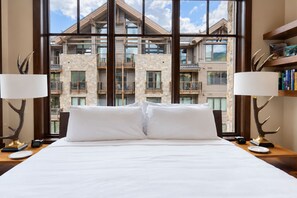 Ein Hotelzimmer mit einem großen Bett, zwei Nachttischlampen und Blick auf ein Mehrfamilienhaus mit Balkonen.