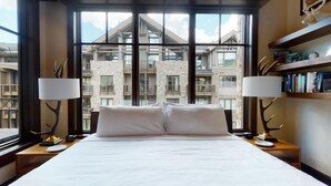 Habitación de hotel con una cama grande, dos lámparas de noche, una mesita de noche y vistas al exterior del edificio a través de amplias ventanas.