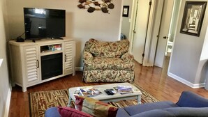 A living room with a television on a cabinet, a floral-patterned chair, a coffee table with magazines, and a blue sofa.