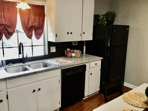 Una cocina con armarios blancos, un refrigerador negro, un lavavajillas y una ventana con cortinas.