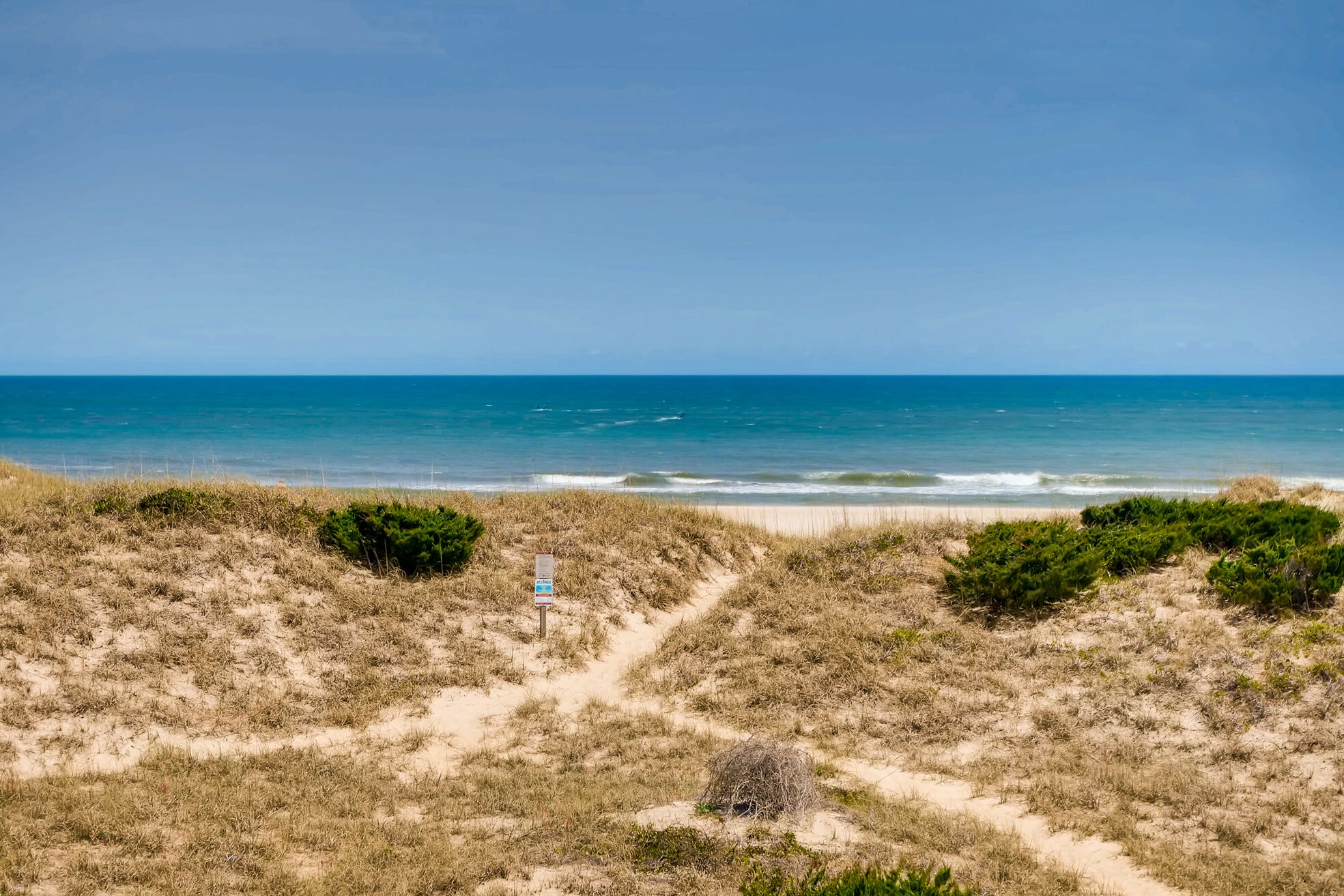 A beach with a clear view of the ocean, sandy dunes, and a signpost.