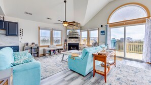 Un salon spacieux avec un grand canapé, une table basse et une cheminée. De nombreuses fenêtres offrent une vue sur la plage et l’océan.
