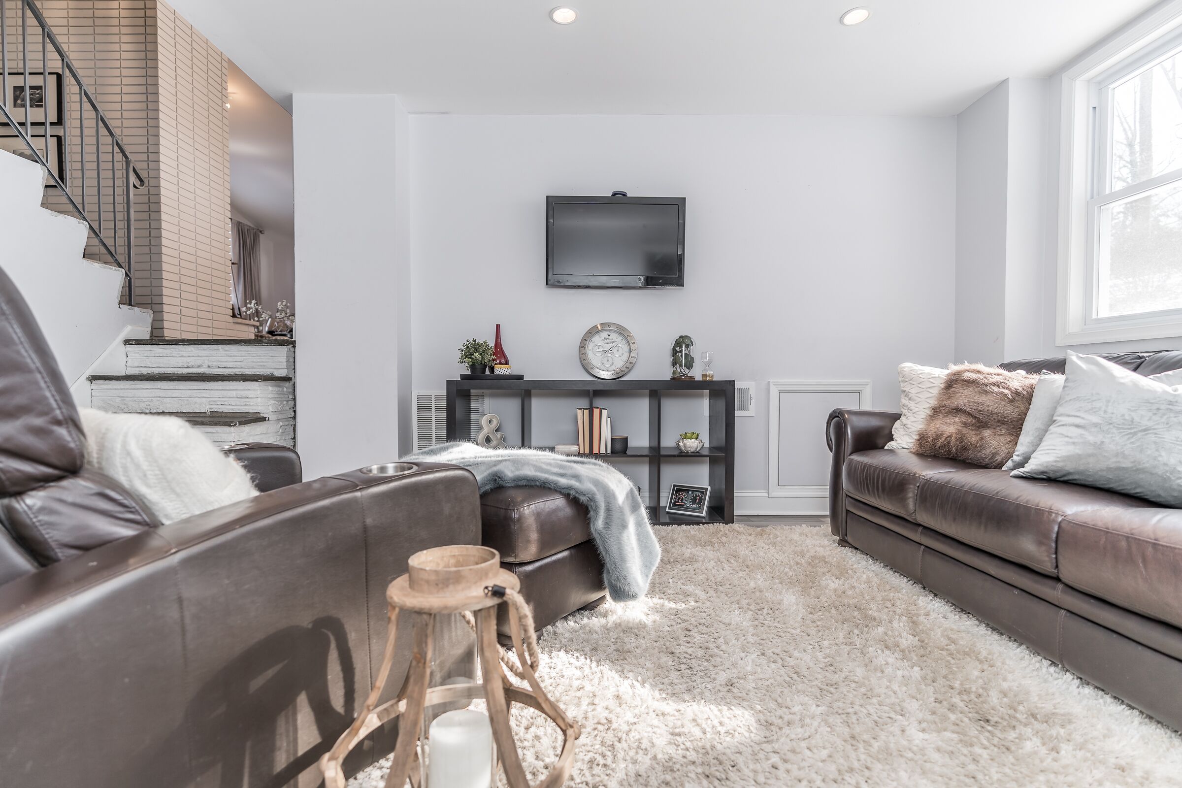 A modern living room with a staircase, leather sofas, a TV, and a bookshelf.