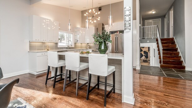 A modern kitchen with white cabinetry, a central island with bar stools, pendant lighting, and a view of a staircase leading to a second floor. - Newly Renovated Mid-Century Home (Newtown Square)