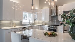 A modern kitchen with white cabinetry, a central island with fruit, pendant lighting, and a large window. - Newly Renovated Mid-Century Home (Newtown Square)