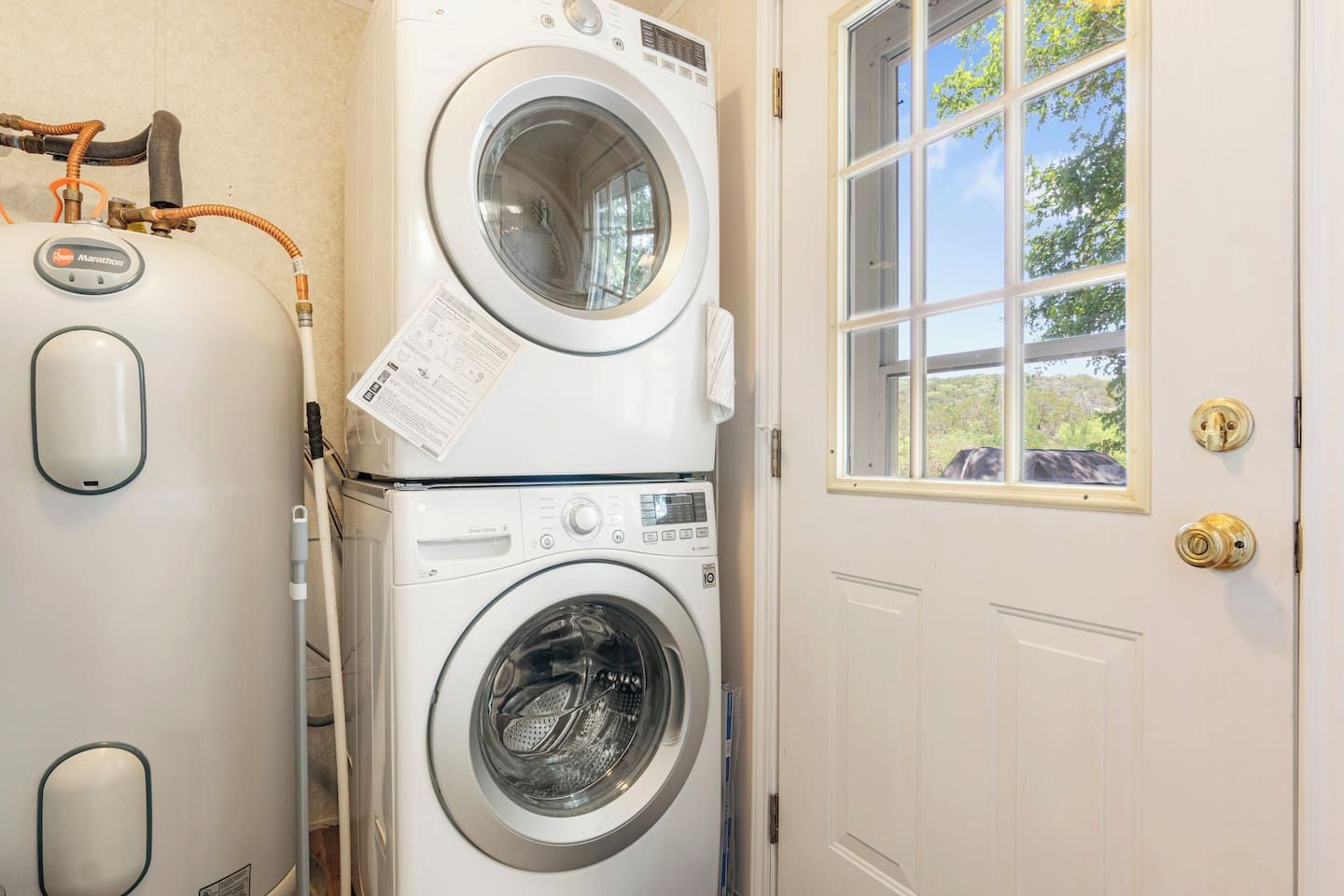 A laundry room with two stacked washing machines, a window with a view of greenery, and a door with a brass handle.